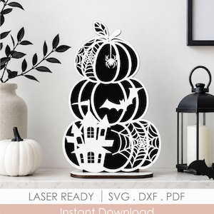 May include: A tiered stack of three pumpkins cut from black and white material. The top pumpkin features a spider web and spider, the middle has a bat silhouette, and the bottom displays a haunted house and spider web. Text reads LASER READY.