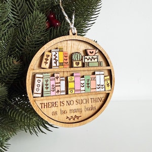 May include: Wooden, round ornament featuring a bookshelf design with colorful books, a cactus, and a teacup. The text "THERE IS NO SUCH THING as too many books" is engraved on the ornament. A white string is attached for hanging.
