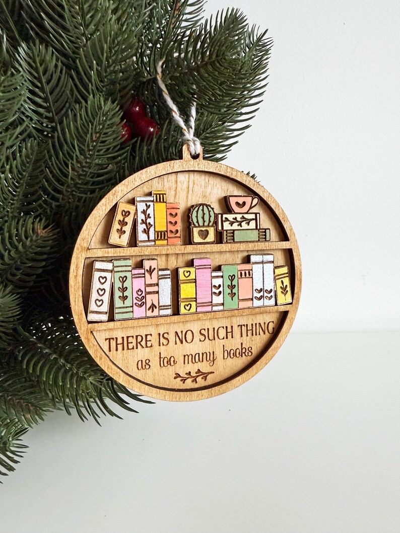 May include: Wooden, round ornament featuring a bookshelf design with colorful books, a cactus, and a teacup. The text "THERE IS NO SUCH THING as too many books" is engraved on the ornament. A white string is attached for hanging.