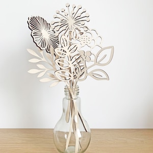 May include: A clear glass vase holds a bouquet of wooden flowers. The flowers are intricately cut from light-colored wood, featuring various shapes like leaves, round blooms, and spiky designs. The vase sits on a light-colored wooden surface against a white background.