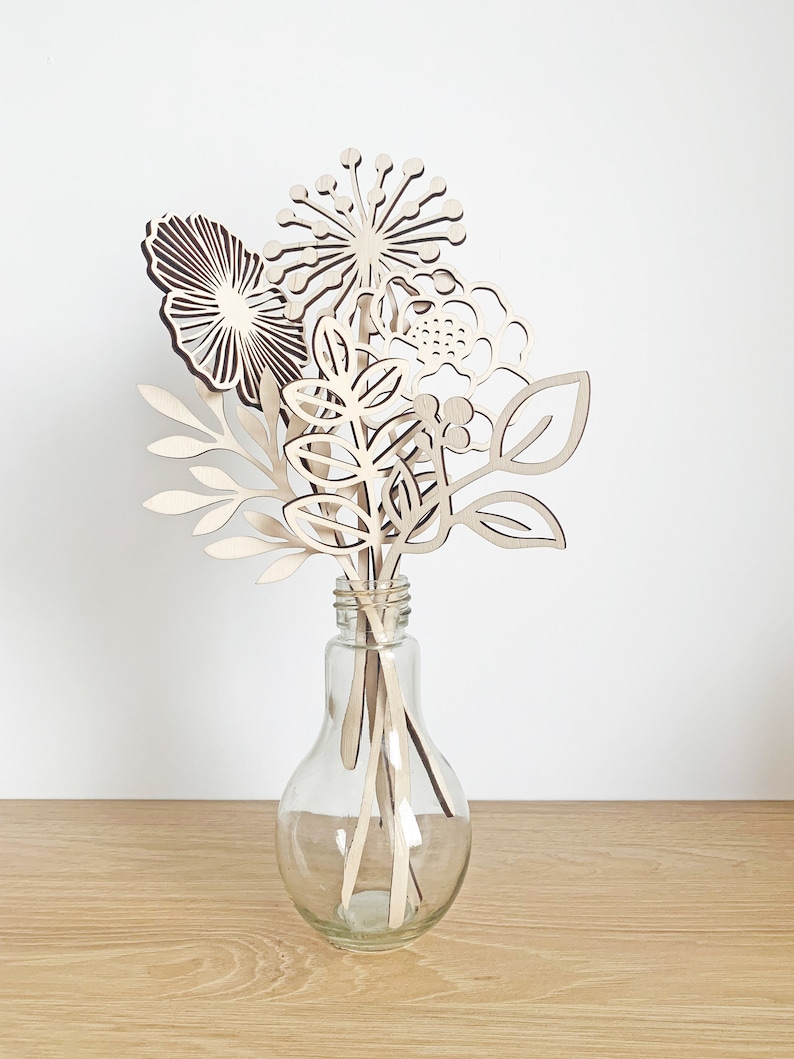 May include: A clear glass vase holds a bouquet of wooden flowers. The flowers are intricately cut from light-colored wood, featuring various shapes like leaves, round blooms, and spiky designs. The vase sits on a light-colored wooden surface against a white background.