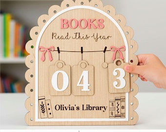 May include: Wooden book tracker with the text "BOOKS Read this Year" in pink. It has three hanging tags with the numbers "04" and "3". The bottom of the tracker says "Olivia's Library". The frame is scalloped and has pink bows.