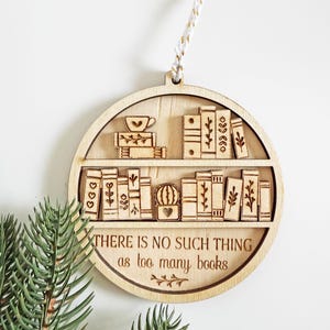May include: Wooden ornament shaped like a bookshelf, featuring engraved books, a teacup, and the text "THERE IS NO SUCH THING as too many books." The ornament is round with a hanging string.