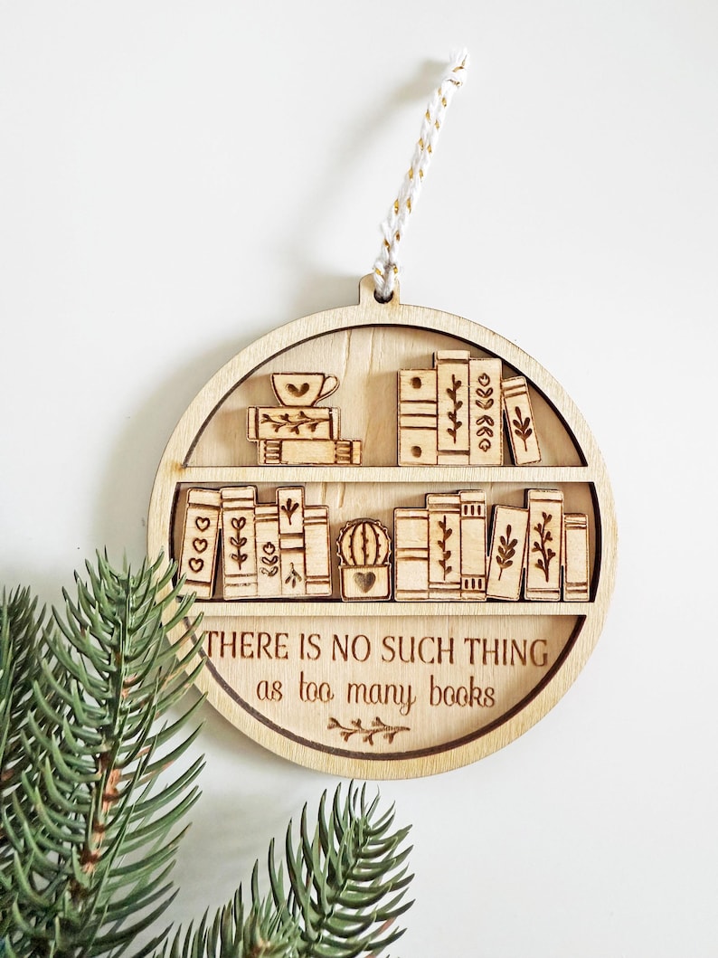 May include: Wooden ornament shaped like a bookshelf, featuring engraved books, a teacup, and the text "THERE IS NO SUCH THING as too many books." The ornament is round with a hanging string.