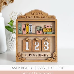 Puede incluir: Expositor de madera con forma de estantería con el texto "BOOKS Read This Year". La estantería presenta libros en miniatura, un cactus y una taza de té. Debajo hay tres etiquetas numeradas y una placa con el nombre "ALLISON'S LIBRARY".