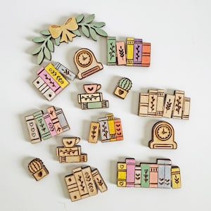 May include: A collection of wooden book-themed embellishments, including stacks of books in various colors, a clock, a bow, a sprig of leaves, a cactus, and a teacup. The embellishments are made of light-colored wood with etched details and painted accents.