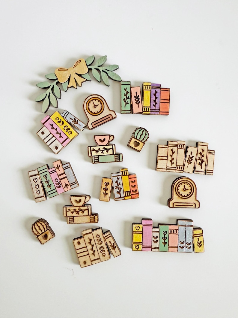 May include: A collection of wooden book-themed embellishments, including stacks of books in various colors, a clock, a bow, a sprig of leaves, a cactus, and a teacup. The embellishments are made of light-colored wood with etched details and painted accents.