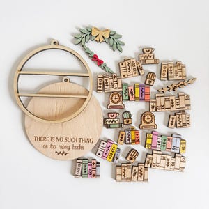 May include: Wooden bookshelf-themed ornaments and a round shelf with the text "THERE IS NO SUCH THING as too many books." The ornaments include miniature books, clocks, and a wreath with a bow. The color palette includes natural wood, green, pink, yellow, and blue.