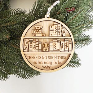 May include: Wooden Christmas ornament with a bookshelf design. The ornament features a circular shape with laser-cut details of books, a cactus, and a teacup. The text "THERE IS NO SUCH THING as too many books" is also included.