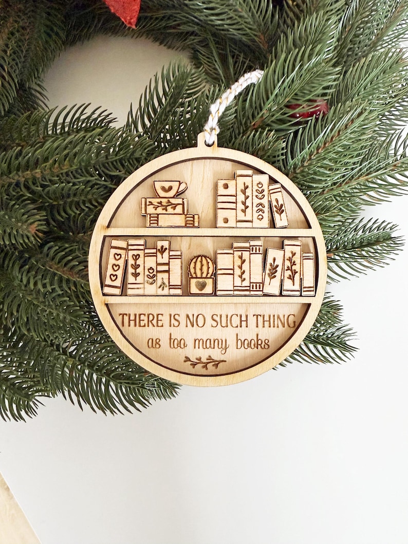 May include: Wooden Christmas ornament with a bookshelf design. The ornament features a circular shape with laser-cut details of books, a cactus, and a teacup. The text "THERE IS NO SUCH THING as too many books" is also included.