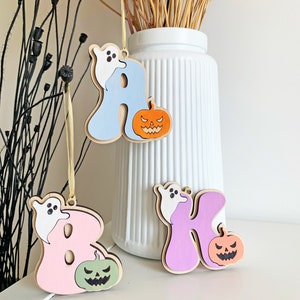 May include: Three wooden letter ornaments hanging by thin yellow strings. The letters A, B, and K are painted in light blue, pink, and purple. Each letter features a small white ghost and a carved jack-o'-lantern pumpkin in orange, green, or peach.