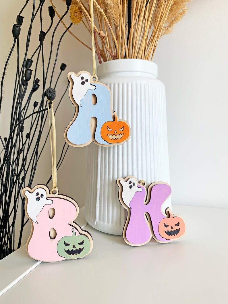 May include: Three wooden letter ornaments hanging by thin yellow strings. The letters A, B, and K are painted in light blue, pink, and purple. Each letter features a small white ghost and a carved jack-o'-lantern pumpkin in orange, green, or peach.