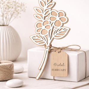 Puede incluir: Un ramo floral de madera con detalles blancos, atado a una caja de regalo blanca con hilo. Una etiqueta de madera dice "Violet February". El ramo presenta flores y hojas.