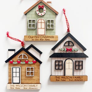 May include: Three wooden house-shaped Christmas ornaments with red hanging strings. Each ornament features a different design, including a green house with a wreath, a brown house with a garland, and a white house with a bow. Text includes "Our First Christmas" and "The Wilson Family."