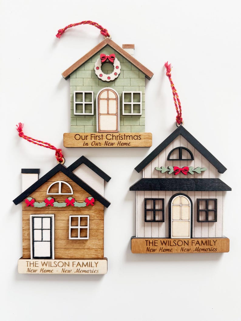 May include: Three wooden house-shaped Christmas ornaments with red hanging strings. Each ornament features a different design, including a green house with a wreath, a brown house with a garland, and a white house with a bow. Text includes "Our First Christmas" and "The Wilson Family."