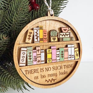 May include: Wooden, round Christmas ornament featuring a bookshelf design. The ornament has two shelves with colorful, miniature books, a cactus, and a teacup. The text "THERE IS NO SUCH THING as too many books" is engraved on the bottom. A string is attached for hanging.