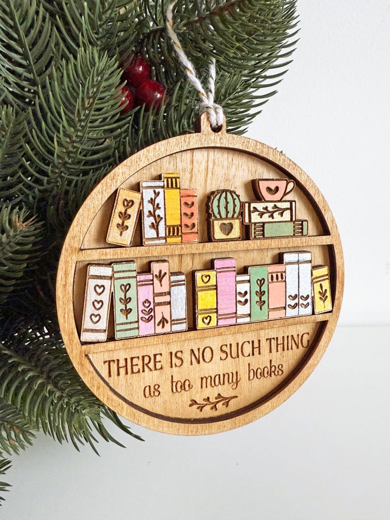 May include: Wooden, round Christmas ornament featuring a bookshelf design. The ornament has two shelves with colorful, miniature books, a cactus, and a teacup. The text "THERE IS NO SUCH THING as too many books" is engraved on the bottom. A string is attached for hanging.