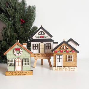 May include: Three wooden house ornaments with Christmas themes. One is green with a wreath, another is white, and the third is brown. Each has a small sign with text. A Christmas tree branch is in the background.
