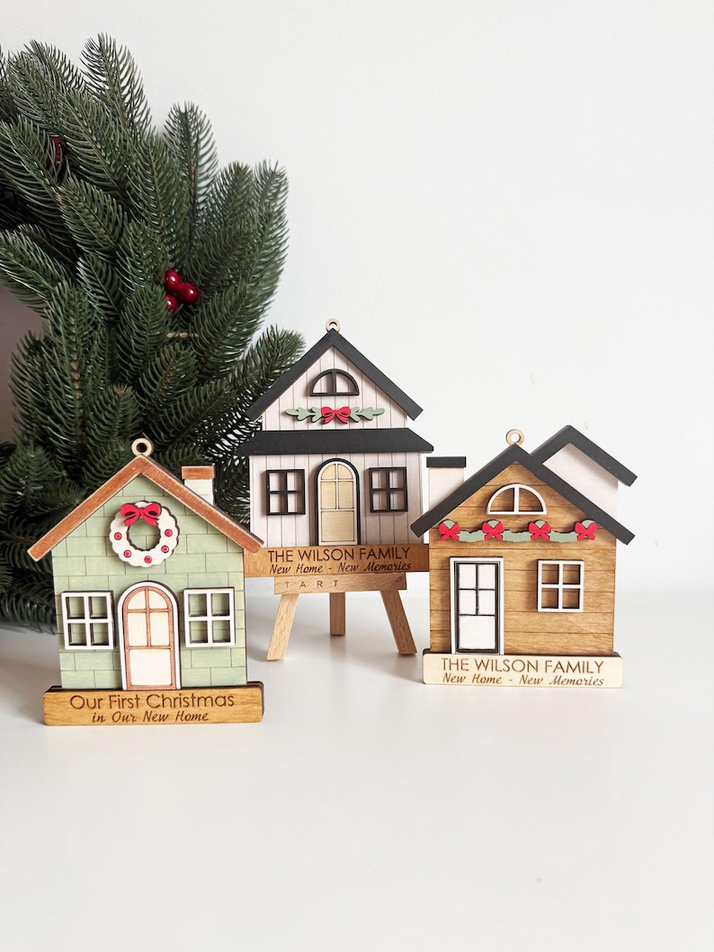 May include: Three wooden house ornaments with Christmas themes. One is green with a wreath, another is white, and the third is brown. Each has a small sign with text. A Christmas tree branch is in the background.