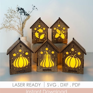 May include: Five small, dark brown wooden house-shaped lanterns with cut-out designs of pumpkins, acorns, and oak leaves. Each house features a warm yellow light glowing from within. Text at the bottom reads: LASER READY | SVG . DXF . PDF