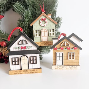 May include: Three wooden house-shaped Christmas ornaments. One is white with black trim, another is light green, and the third is brown. Each has a red string for hanging and the text "THE WILSON FAMILY New Home - New Memories".