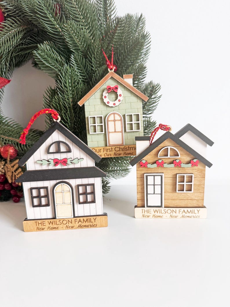 May include: Three wooden house-shaped Christmas ornaments. One is white with black trim, another is light green, and the third is brown. Each has a red string for hanging and the text "THE WILSON FAMILY New Home - New Memories".