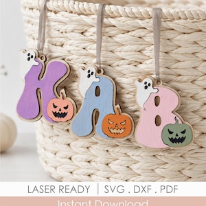 May include: Three wooden hanging ornaments shaped as the letters K, A, and B. Each letter features a small white ghost and a carved pumpkin with a jagged face. The letters are painted in purple, light blue, and pink, suspended by grey ribbons.
