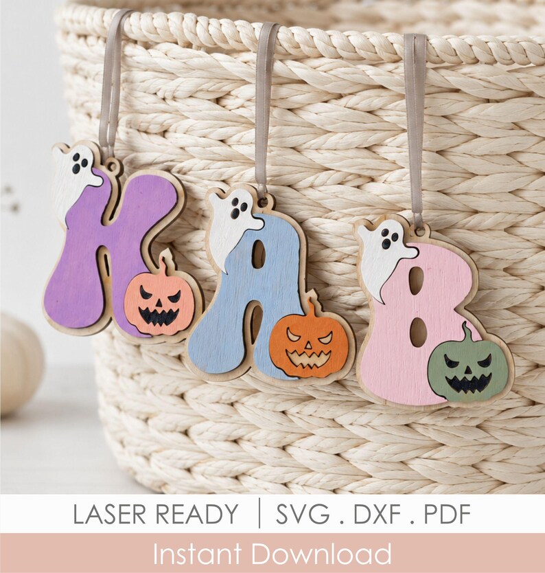 May include: Three wooden hanging ornaments shaped as the letters K, A, and B. Each letter features a small white ghost and a carved pumpkin with a jagged face. The letters are painted in purple, light blue, and pink, suspended by grey ribbons.