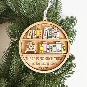 May include: Wooden ornament featuring a bookshelf design with colorful books and a clock. The text "THERE IS NO SUCH THING as too many books" is engraved on the ornament. The ornament is hanging on a green Christmas tree.