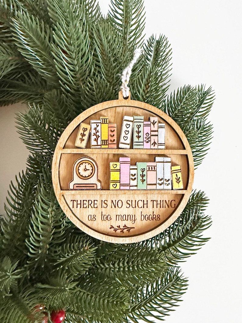 May include: Wooden ornament featuring a bookshelf design with colorful books and a clock. The text "THERE IS NO SUCH THING as too many books" is engraved on the ornament. The ornament is hanging on a green Christmas tree.