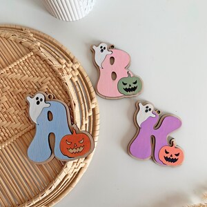 May include: Three wooden letter ornaments shaped as A, B, and K. The letter A is light blue with a white ghost and orange jack-o'-lantern. The letter B is pink with a white ghost and green jack-o'-lantern. The letter K is purple with a white ghost and orange jack-o'-lantern.