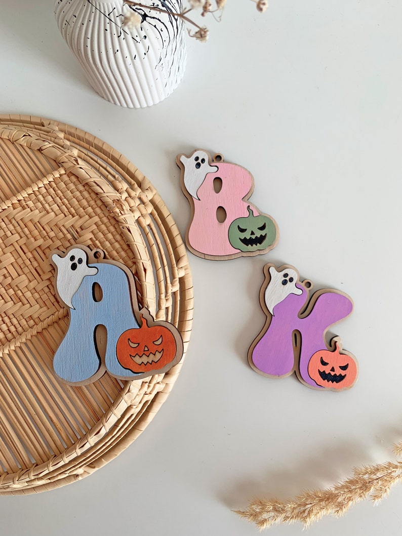 May include: Three wooden letter ornaments shaped as A, B, and K. The letter A is light blue with a white ghost and orange jack-o'-lantern. The letter B is pink with a white ghost and green jack-o'-lantern. The letter K is purple with a white ghost and orange jack-o'-lantern.