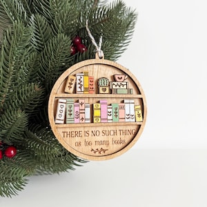 May include: Wooden, round Christmas ornament featuring a bookshelf design with colorful books and a cactus. The text "THERE IS NO SUCH THING as too many books" is engraved on the ornament. A string is attached for hanging.