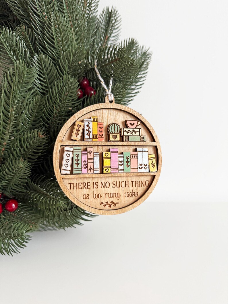 May include: Wooden, round Christmas ornament featuring a bookshelf design with colorful books and a cactus. The text "THERE IS NO SUCH THING as too many books" is engraved on the ornament. A string is attached for hanging.