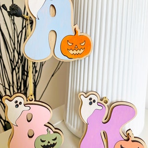 May include: Three wooden hanging ornaments shaped like the letters A, B, and K. Each letter features a white ghost and a jack-o'-lantern. The letter A is light blue with an orange pumpkin, B is pink with a sage green pumpkin, and K is purple with an orange pumpkin.