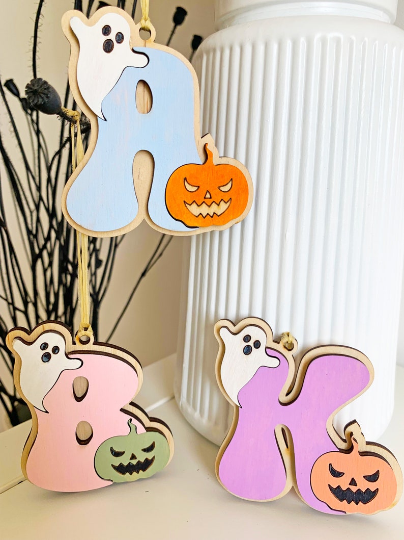 May include: Three wooden hanging ornaments shaped like the letters A, B, and K. Each letter features a white ghost and a jack-o'-lantern. The letter A is light blue with an orange pumpkin, B is pink with a sage green pumpkin, and K is purple with an orange pumpkin.