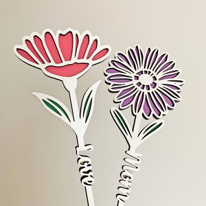 May include: Two white wooden flowers with green leaves and the words "Mom" and "Love" written on the stems. The flowers are pink and purple.