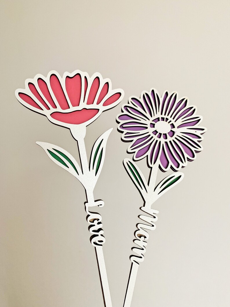 May include: Two white wooden flowers with green leaves and the words "Mom" and "Love" written on the stems. The flowers are pink and purple.