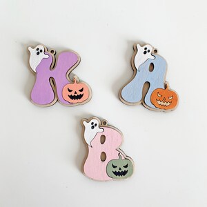 May include: Three wooden letter ornaments shaped as K, A, and B. The K is purple, the A is light blue, and the B is light pink. Each letter features a small white ghost attached to the top and a carved jack-o'-lantern pumpkin at the base.