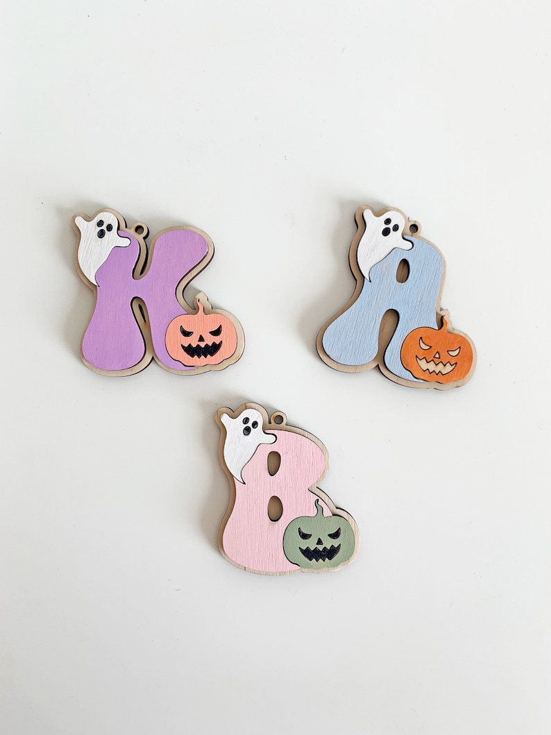 May include: Three wooden letter ornaments shaped as K, A, and B. The K is purple, the A is light blue, and the B is light pink. Each letter features a small white ghost attached to the top and a carved jack-o'-lantern pumpkin at the base.