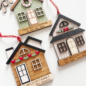 May include: Wooden house-shaped Christmas ornaments with red hanging strings. One is green with a wreath, another is brown with a garland, and the third is white with a garland. Each has text: "Our First Christmas in Our New Home" or "The Wilson Family New Home - New Memories."