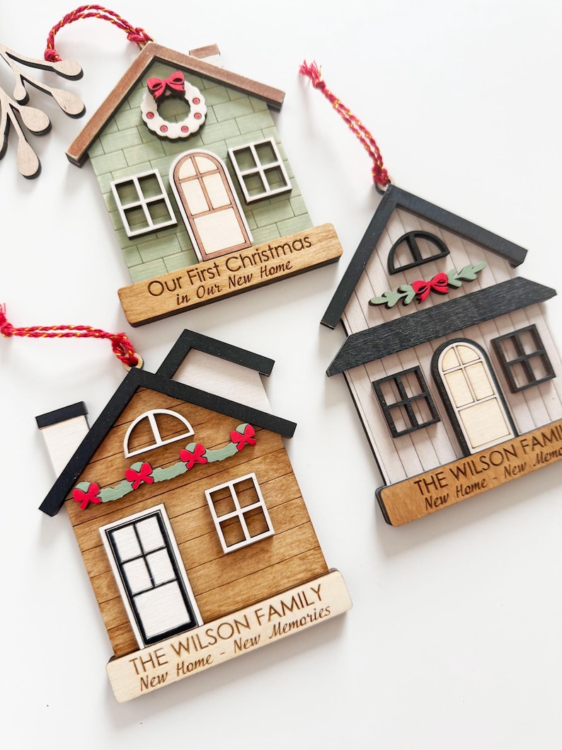 May include: Wooden house-shaped Christmas ornaments with red hanging strings. One is green with a wreath, another is brown with a garland, and the third is white with a garland. Each has text: "Our First Christmas in Our New Home" or "The Wilson Family New Home - New Memories."