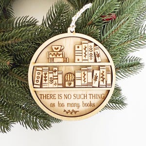 May include: A wooden Christmas ornament featuring a bookshelf design with books, a teacup, and a small cactus. The ornament has the text "THERE IS NO SUCH THING as too many books" and is hanging from a white string. The ornament is set against a backdrop of green pine needles.
