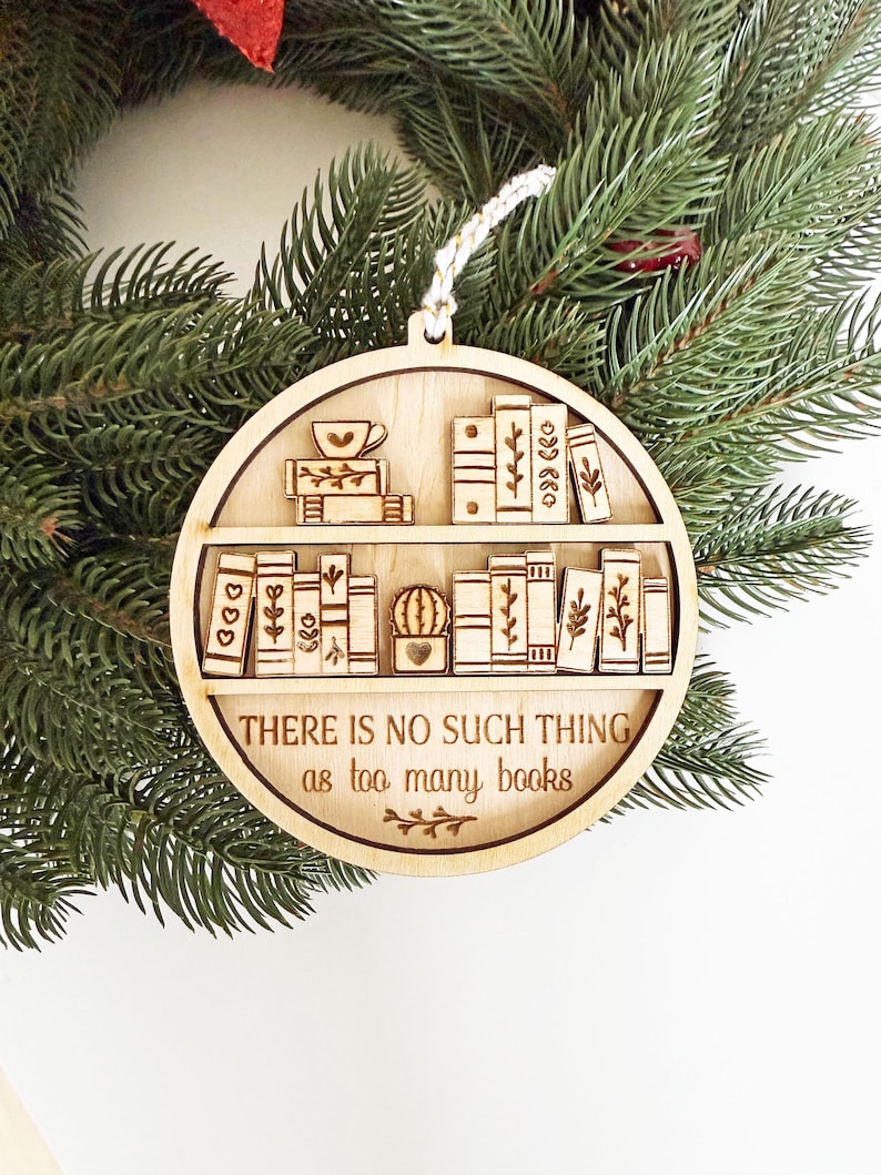 May include: A wooden Christmas ornament featuring a bookshelf design with books, a teacup, and a small cactus. The ornament has the text "THERE IS NO SUCH THING as too many books" and is hanging from a white string. The ornament is set against a backdrop of green pine needles.