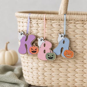 May include: Three wooden letter ornaments spelling KBA, each painted in pastel purple, pink, and blue. Each letter features a small white ghost and a carved jack-o'-lantern pumpkin. The letters hang from matching pastel ribbons on a woven basket.
