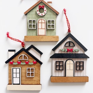 May include: Three wooden house ornaments with red hanging strings. The houses have different designs, including green, brown, and white exteriors, with details like wreaths, bows, and window and door frames. These ornaments are perfect for holiday decor.