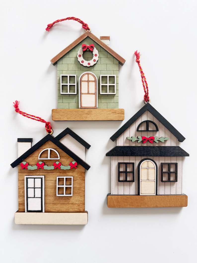 May include: Three wooden house ornaments with red hanging strings. The houses have different designs, including green, brown, and white exteriors, with details like wreaths, bows, and window and door frames. These ornaments are perfect for holiday decor.