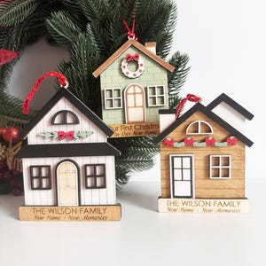 May include: Three wooden house ornaments with red hanging strings. The houses are painted white, green, and brown, with black and white accents. Each ornament has a small sign with text. One says "Our First Christmas In Our New Home."