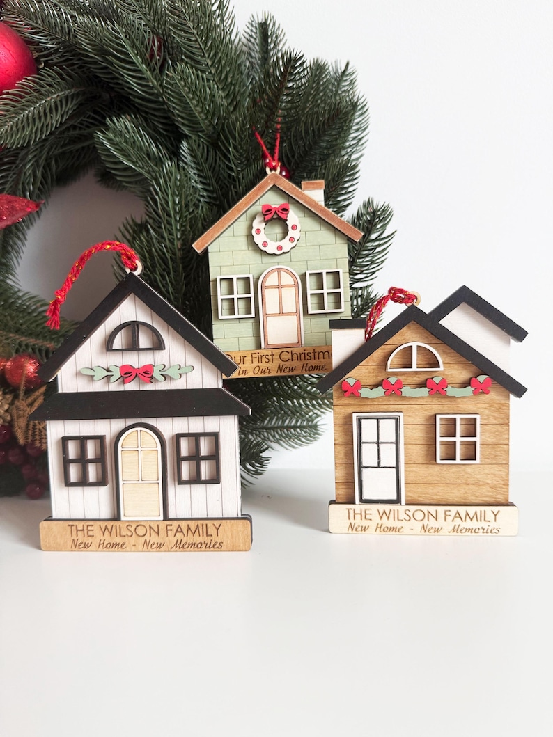May include: Three wooden house ornaments with red hanging strings. The houses are painted white, green, and brown, with black and white accents. Each ornament has a small sign with text. One says "Our First Christmas In Our New Home."