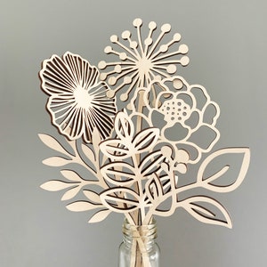 May include: A bouquet of wooden flowers in a clear glass bottle. The flowers are cut from light-colored wood and feature various floral and leaf designs. The arrangement includes a large, open flower, a spiky bloom, and several leafy stems.